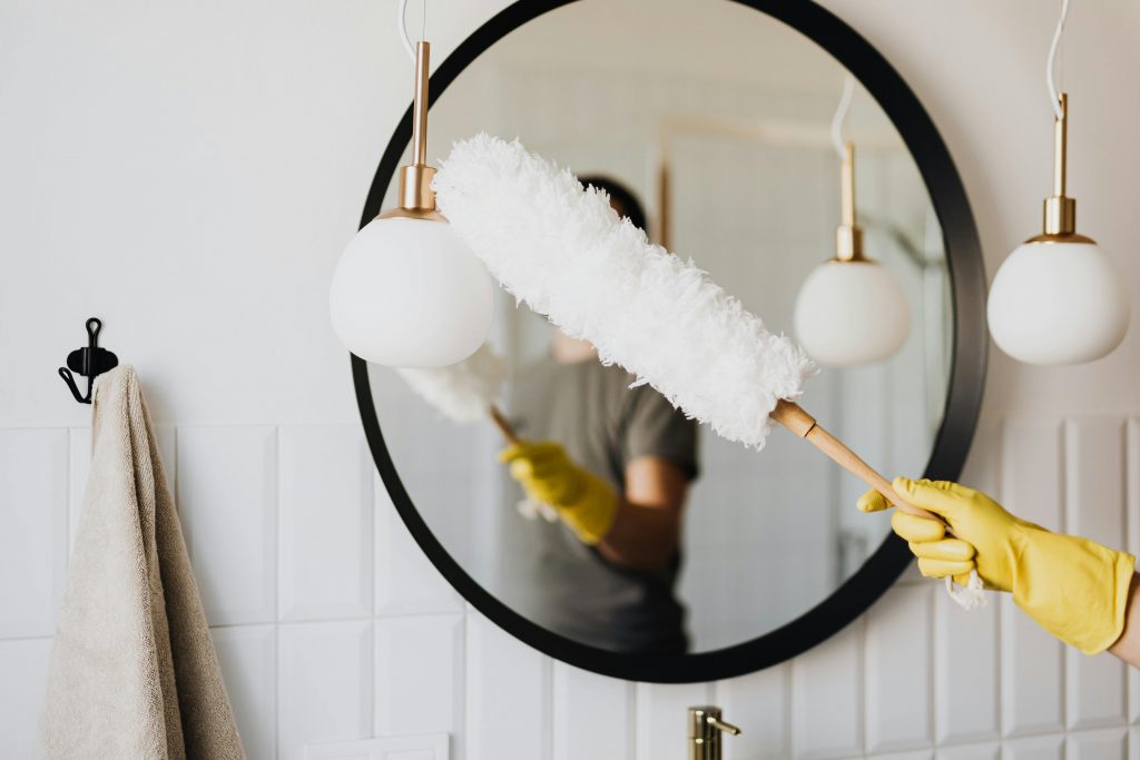Crop anonymous female housewife in rubber gloves dusting modern lamp with soft brush against mirror while cleaning stylish bathroom