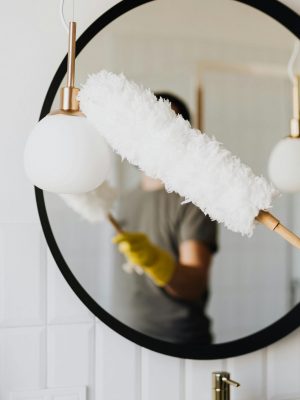 Crop anonymous female housewife in rubber gloves dusting modern lamp with soft brush against mirror while cleaning stylish bathroom